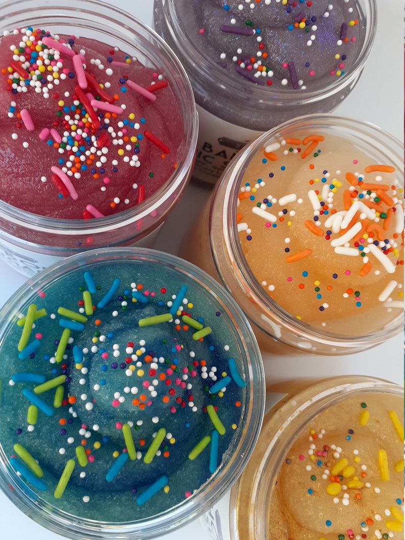 May include: Four clear plastic jars filled with colorful, textured slime. Each jar features a distinct color: deep pink, dark purple, bright orange, and teal blue. The surface of each slime is topped with a variety of small, multi-colored sprinkles.