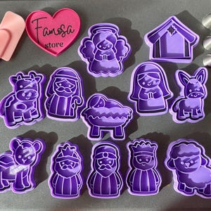 May include: A collection of purple plastic cookie cutters in various shapes, including an angel, a house, a cow, a man with a staff, a baby in a manger, a woman, a donkey, a lamb, and three kings. A pink heart-shaped object with the text "Famosa store" is also present.