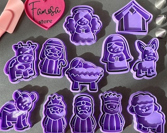May include: A collection of purple plastic cookie cutters in various shapes, including an angel, a house, a cow, a man with a staff, a baby in a manger, a woman, a donkey, a lamb, and three kings. A pink heart-shaped object with the text "Famosa store" is also present.