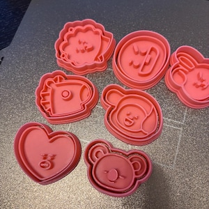 May include: A collection of seven coral-colored plastic cookie cutters featuring various cartoon character faces and shapes, including a heart, a bear, and other stylized figures with raised internal details for imprinting dough designs.