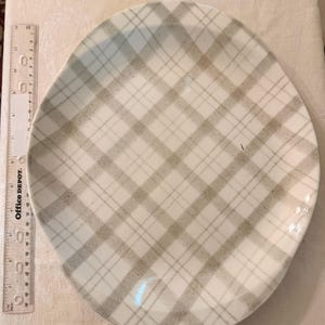 May include: A round ceramic plate featuring a beige and white plaid pattern. A clear plastic ruler is positioned vertically along the left side of the plate, showing the item measures approximately 10.5 inches in diameter at its widest point.