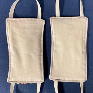May include: Two light gray fabric face masks with white elastic ear loops are displayed against a dark blue background. The masks have a rectangular shape with rounded corners and are sewn with a contrasting gray thread.