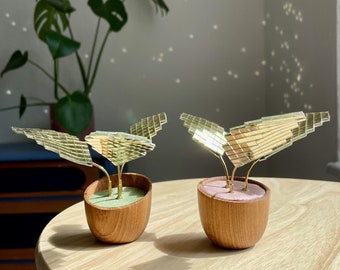 May include: Two small potted plants with gold mirrored leaves. The plants are in wooden pots with a green and pink interior. The pots are sitting on a light wood table.