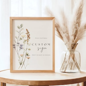 May include: A printable sign with a floral design and the text "FULLY EDITABLE CUSTOM sign PRINT AS MANY AS NEEDED". The sign is framed in a light wood frame and is sitting on a light wood table.