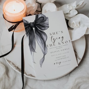 May include: A bridal shower invitation with a watercolor-style black bow illustration. The invitation reads "She's Tying the Knot" with event details including the honoree's name, date, time, and location. A lit candle and black ribbon are also in the image.