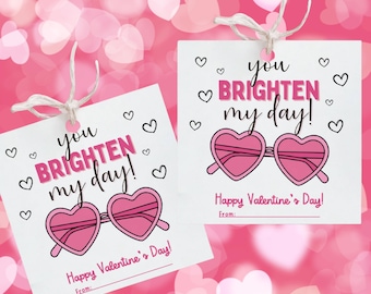 May include: Two white gift tags with the text "you brighten my day!" and "Happy Valentine's Day!" in pink. Each tag features a pair of pink heart-shaped sunglasses and small heart illustrations. The tags are tied with twine.