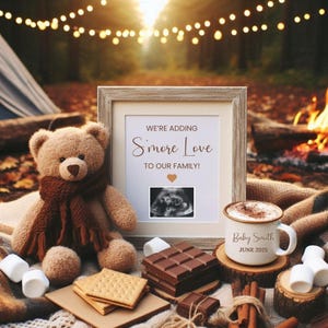 Könnte beinhalten: Eine gerahmte Ankündigung mit dem Text "We're adding S'more Love to our family!" mit einem Herz und einem Ultraschallbild im Inneren. Ein Teddybär mit einem Schal sitzt neben dem Rahmen. Eine Tasse mit dem Text "Baby Smith June 2025" steht auf einem Holzstamm mit einem Teller Graham-Cracker und Schokoladentafeln.