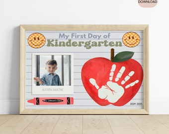 May include: A printable first day of kindergarten sign featuring a red apple, a handprint, a smiley face, and a photo frame. The sign reads "My First Day of Kindergarten" and includes the text "Aiden Baker" and "Mrs. Johnson's Class".