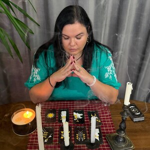 May include: A person with long dark hair wearing a green embroidered blouse sits with hands pressed together. On a red plaid cloth, eight tarot cards are arranged in two rows, accompanied by three white candles and a lit candle in a tin.