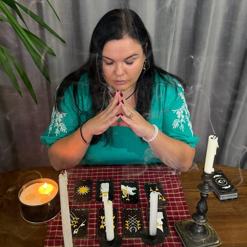 May include: A person with long dark hair wearing a green embroidered blouse sits with hands pressed together. On a red plaid cloth, eight tarot cards are arranged in two rows, accompanied by three white candles and a lit candle in a tin.