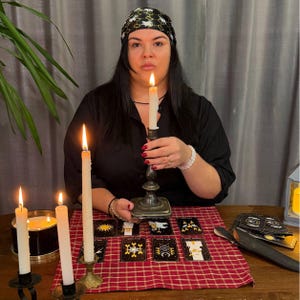 May include: A black headscarf with a white bee pattern, a black shirt, and a silver candlestick holding a lit white taper candle. On the table are eight tarot cards, a red and white checkered cloth, and two additional lit white candles.