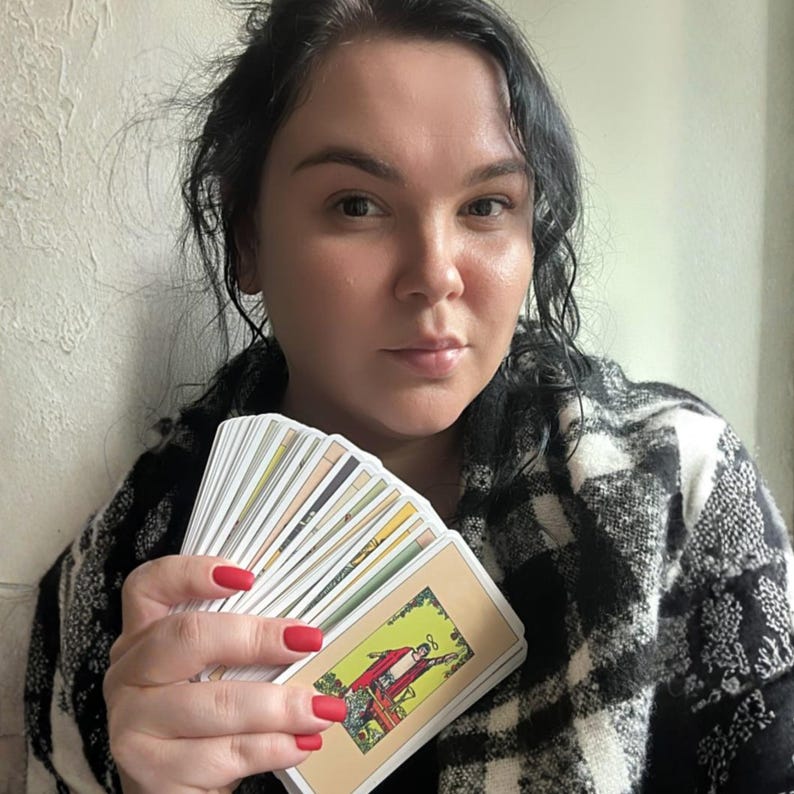May include: A deck of tarot cards held in a hand with red nail polish. The front card features a colorful illustration of a figure in a red robe standing in a green landscape. The cards have white borders and are fanned out to show multiple edges.