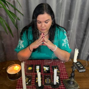 May include: A person with long dark hair wearing a green embroidered blouse sits with hands pressed together. On a red plaid cloth, eight tarot cards are arranged in two rows, accompanied by three white candles and a lit candle in a tin.