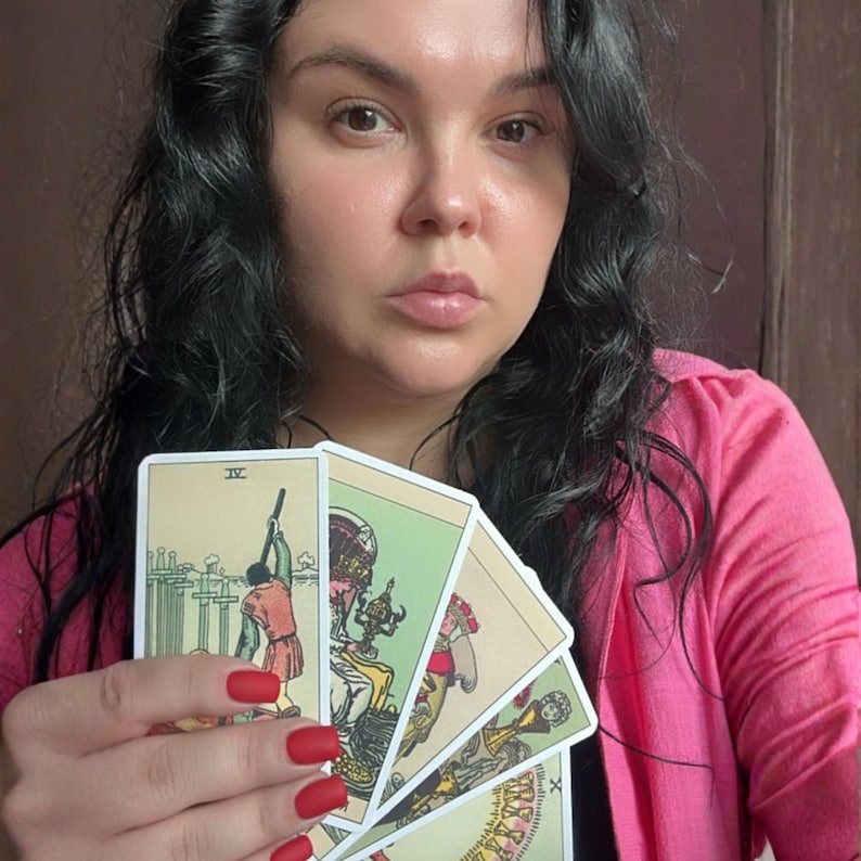 May include: A set of tarot cards held in a hand with matte red fingernails. The cards feature illustrated scenes including a figure with swords, a seated figure with a chalice, and other figures in muted tones of green, tan, and red.