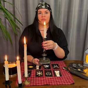 May include: A black headscarf with a white bee pattern, a black shirt, and a silver candlestick holding a lit white taper candle. On the table are eight tarot cards, a red and white checkered cloth, and two additional lit white candles.