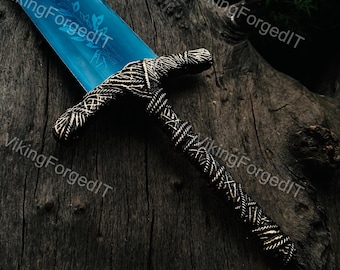 Könnte beinhalten: Nahaufnahme eines dekorativen Schwertes mit einer blauen Klinge und einem detaillierten, gemusterten Griff. Die Klinge weist ein florales Design auf. Der Griff ist aufwendig mit einer dunklen Metalloberfläche gewebt. Das Schwert wird auf einer dunklen, strukturierten Oberfläche präsentiert.