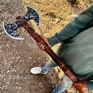Könnte beinhalten: Eine Doppelaxt mit einem Holzstiel, der in braunes Leder gewickelt ist. Die Axt hat eine schwarze Metalloberfläche und wird von einer Person in blauer Jeans und einem grauen Sweatshirt gehalten. Der Text "Viking Forged IT" ist in den Stiel graviert.