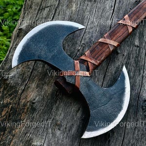 Könnte beinhalten: Eine Doppelaxt mit dunkler, strukturierter Klinge und einem Holzgriff, der mit Leder umwickelt ist. Der Axtkopf hat eine gebogene Form mit einer scharfen Kante. Der Griff hat eine dunkelbraune Farbe. Die Axt wird auf einer verwitterten Holzoberfläche präsentiert.