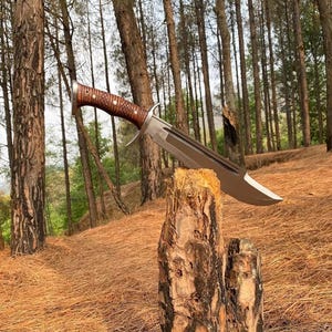 May include: A large, silver-colored knife with a brown wooden handle is resting on a tree stump in a forest setting. The knife has a curved blade and a serrated edge.