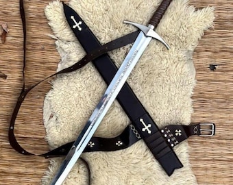May include: A medieval sword with a silver blade and brown handle, resting on a white fur-like surface. The sword is in a black sheath with a brown leather belt, adorned with cross symbols.