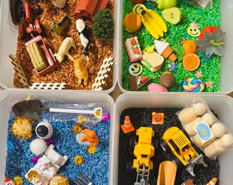 May include: Four rectangular plastic bins filled with sensory materials and small toys. One bin contains brown rice with farm animal figures, a metal bucket, and a red barn. Another holds green rice with assorted plastic food items. A third features blue rice with space-themed figures and a rocket. The final bin contains black rice with construction vehicles, traffic cones, and wooden blocks.