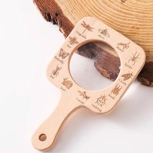 May include: A wooden magnifying glass with a handle and a round magnifying lens. The frame is decorated with images of insects and the names of the insects are printed around the frame: Dragonfly, Moth, Mantis, Spider, Cicada, Beetle, Ladybug, Locust, Snail, Ant, Butterfly, Bee.