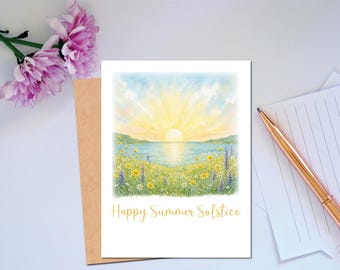 Op de afbeelding: Een wenskaart met een aquarel schilderij van een zonsopgang boven een meer, met velden gele en paarse wilde bloemen. De tekst "Happy Summer Solstice" is in gouden letters geschreven. Een bruine envelop en een pen liggen naast de kaart.