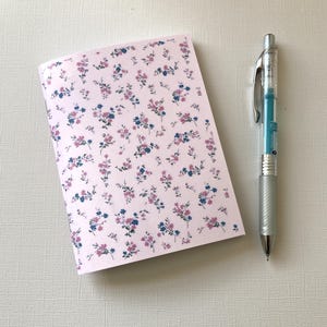 May include: A small notebook with a pink floral pattern featuring blue and purple flowers. A silver pen with a blue ink cartridge is next to the notebook.