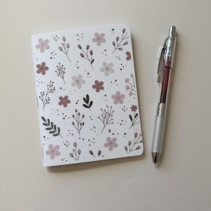 Peut inclure: Un carnet blanc orné d'un motif répétitif de petites fleurs roses, de feuilles vert foncé et de fines tiges brunes, accompagné d'un stylo rétractable argenté avec une cartouche d'encre rouge foncé et une zone de préhension métallique texturée.