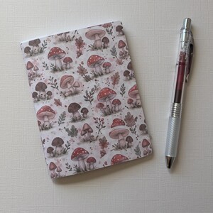 Puede incluir: Un cuaderno rectangular con un patrón repetitivo de varios hongos, hojas pequeñas y bayas en tonos rojos, marrones y verdes sobre un fondo blanco, colocado junto a un bolígrafo retráctil transparente con tinta roja oscura.