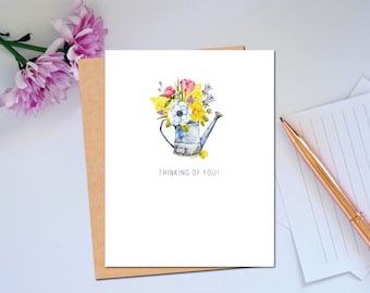 Puede incluir: Una tarjeta blanca con una ilustración de acuarela de un ramo de flores en una regadera azul. El texto "Thinking of you!" está impreso debajo de la imagen.