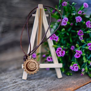 Könnte beinhalten: Eine Holz-Halskette mit einem Sonnen- und Mond-Design. Der Anhänger hängt an einem braunen Lederband und wird auf einem Holzständer präsentiert. Der Hintergrund ist ein verschwommenes Bild von lila Blumen.
