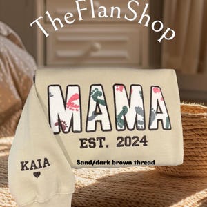 K&ouml;nnte beinhalten: Ein beiger Pullover mit dem Wort "MAMA" in wei&szlig;en Buchstaben mit bunten Fu&szlig;abdr&uuml;cken. Der Text "EST. 2024" befindet sich unter dem Wort "MAMA". Der Text "Sand/dark brown thread" befindet sich unter dem Text "EST. 2024". Der Text "KAIA" befindet sich auf dem &Auml;rmel des Pullovers mit einem kleinen Herzen darunter.