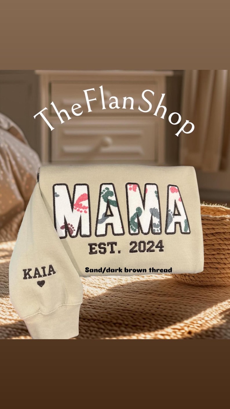 K&ouml;nnte beinhalten: Ein beiger Pullover mit dem Wort "MAMA" in wei&szlig;en Buchstaben mit bunten Fu&szlig;abdr&uuml;cken. Der Text "EST. 2024" befindet sich unter dem Wort "MAMA". Der Text "Sand/dark brown thread" befindet sich unter dem Text "EST. 2024". Der Text "KAIA" befindet sich auf dem &Auml;rmel des Pullovers mit einem kleinen Herzen darunter.