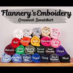 K&ouml;nnte beinhalten: Eine Auswahl an verschiedenen Farben von Crewneck-Sweatshirts, die auf einer Holzoberfl&auml;che aufgerollt und ausgestellt sind. Die Farben umfassen Wei&szlig;, Asche, Sportgrau, Sand, Hellrosa, Rot, Orange, Gold, Heliconia, Sicherheitsrosa, Maroon, Irischgr&uuml;n, Milit&auml;rgr&uuml;n, Waldgr&uuml;n, Dunkelbraun, Lila, Hellblau, Carolina-Blau, Indigo-Blau, K&ouml;nigsblau, Marineblau, Dunkelgrau und Schwarz.
