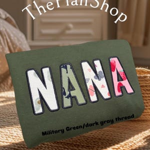 K&ouml;nnte beinhalten: Ein gr&uuml;nes Kissen mit dem Wort "NANA" bestickt in buntem, gemustertem Stoff. Der Text "Milit&auml;rgr&uuml;n/dunkelgrauer Faden" ist unter dem Wort gedruckt.