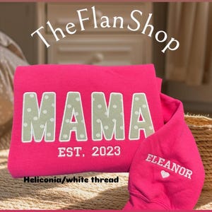 K&ouml;nnte beinhalten: Rosafarbener Pullover mit dem Text "MAMA EST. 2023" in wei&szlig;er Schrift auf der Vorderseite und "ELEANOR" mit einem Herz in wei&szlig;er Schrift auf dem &Auml;rmel. Der Pullover ist aus Heliconia-Stoff gefertigt.