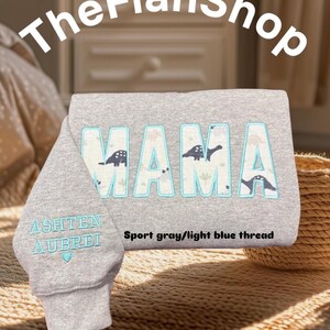 K&ouml;nnte beinhalten: Grauer Sweatshirt mit dem Wort "MAMA" in gro&szlig;en, hellblauen Buchstaben, die mit Schwarz umrandet sind. Die Buchstaben sind mit kleinen Dinosauriersilhouetten verziert. Der Sweatshirt hat eine Tasche am &Auml;rmel mit den Namen "Ashten" und "Aubrei", die in hellblauem Garn gestickt sind. Der Text "Sport grau/hellblauer Faden" ist unter dem Wort "MAMA" gedruckt.