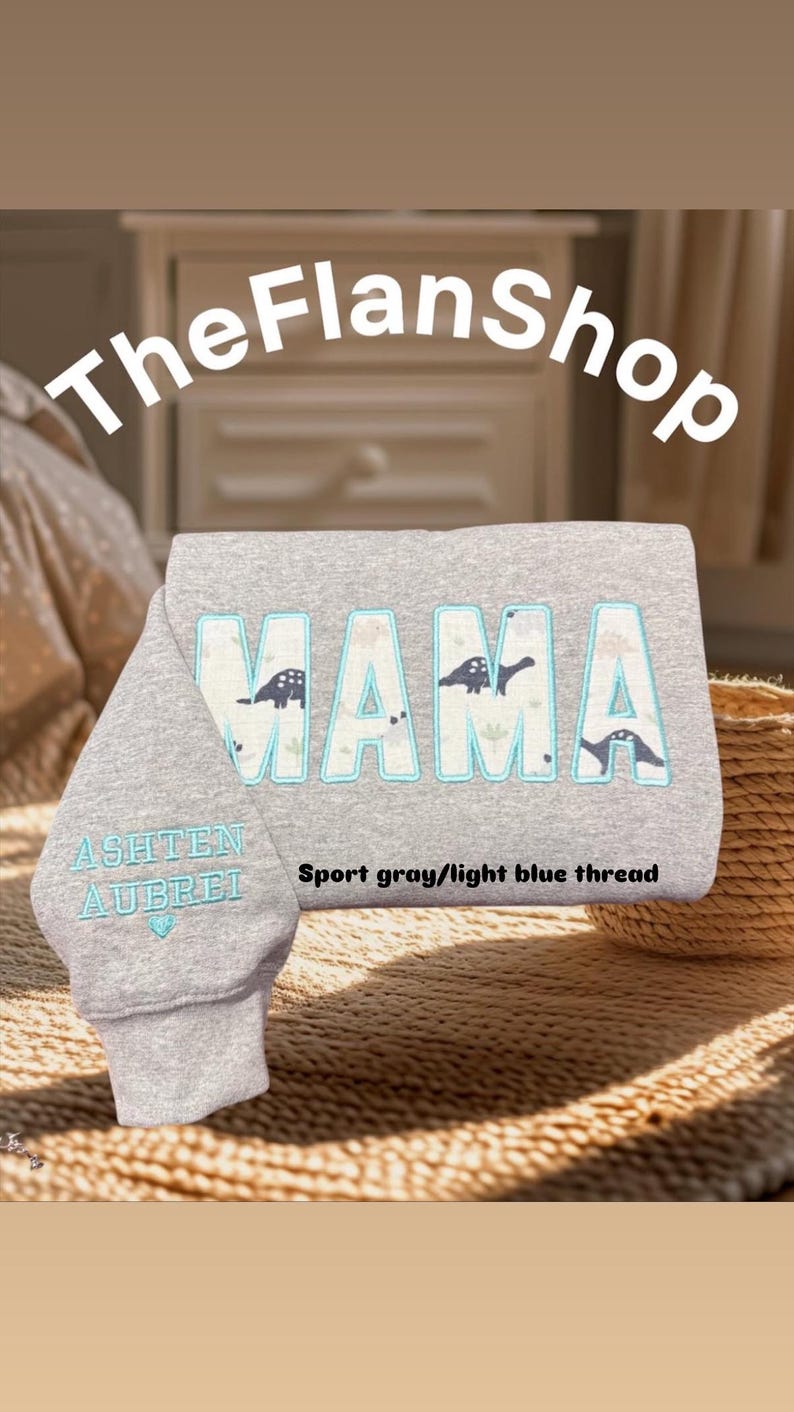 K&ouml;nnte beinhalten: Grauer Sweatshirt mit dem Wort "MAMA" in gro&szlig;en, hellblauen Buchstaben, die mit Schwarz umrandet sind. Die Buchstaben sind mit kleinen Dinosauriersilhouetten verziert. Der Sweatshirt hat eine Tasche am &Auml;rmel mit den Namen "Ashten" und "Aubrei", die in hellblauem Garn gestickt sind. Der Text "Sport grau/hellblauer Faden" ist unter dem Wort "MAMA" gedruckt.