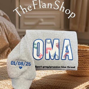 K&ouml;nnte beinhalten: Ein grauer Pullover mit dem Wort "OMA" in blau gestickt, wobei sich innerhalb der Buchstaben ein Muster aus Fu&szlig;abdr&uuml;cken befindet. Das Datum "01/08/25" ist in blau unter dem Wort "OMA" mit einem kleinen Herz gestickt. Der Text "Sport grau/preu&szlig;isch blauer Faden" ist unter dem Datum gedruckt.