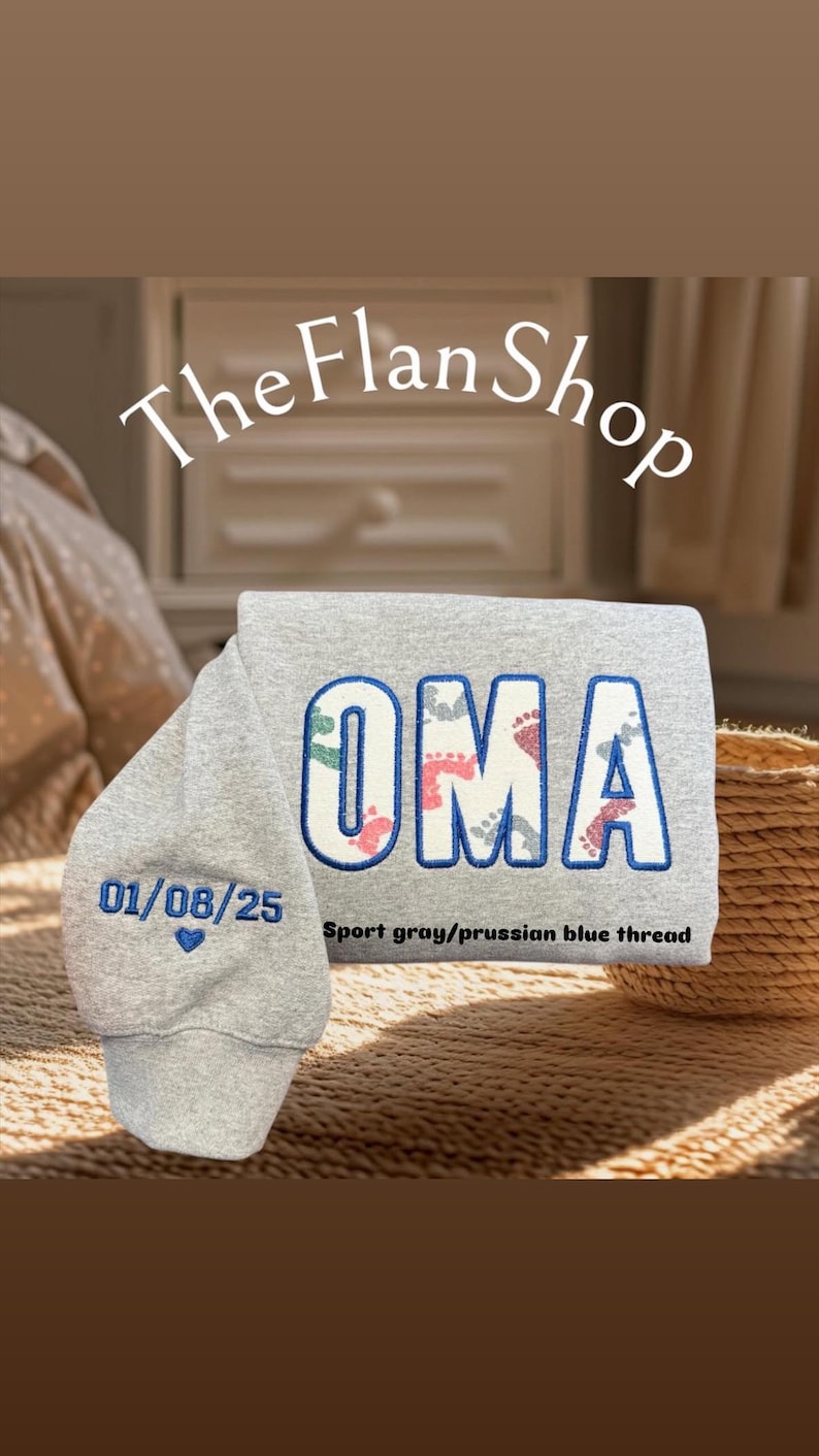K&ouml;nnte beinhalten: Ein grauer Pullover mit dem Wort "OMA" in blau gestickt, wobei sich innerhalb der Buchstaben ein Muster aus Fu&szlig;abdr&uuml;cken befindet. Das Datum "01/08/25" ist in blau unter dem Wort "OMA" mit einem kleinen Herz gestickt. Der Text "Sport grau/preu&szlig;isch blauer Faden" ist unter dem Datum gedruckt.