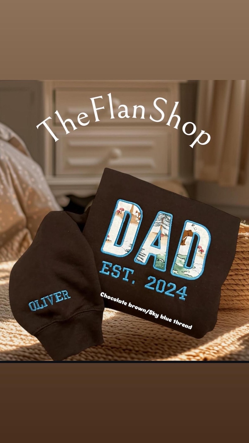 K&ouml;nnte beinhalten: Zwei schwarze Kapuzenpullover mit gesticktem Text. Ein Pullover sagt "DAD EST. 2024" mit einer Waldszene in den Buchstaben. Der andere Pullover sagt "OLIVER". Der Text ist in Himmelblau und Schokoladenbraun gestickt.