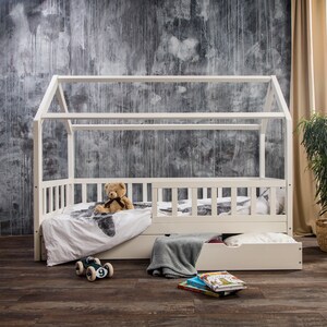 Peut inclure: Cadre de lit en bois blanc en forme de maison avec un matelas blanc et un tiroir de rangement blanc. Un ours en peluche est assis sur le lit et une voiture jouet est sur le sol.