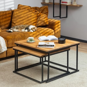 May include: A set of two nesting coffee tables with a black metal frame and a light wood top. The tables are sitting on a light beige rug in front of a mustard yellow sofa.