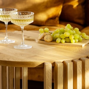 May include: Two glasses of white wine on a wooden table with a bowl of green grapes and cookies. The table has a unique design with vertical wooden slats.