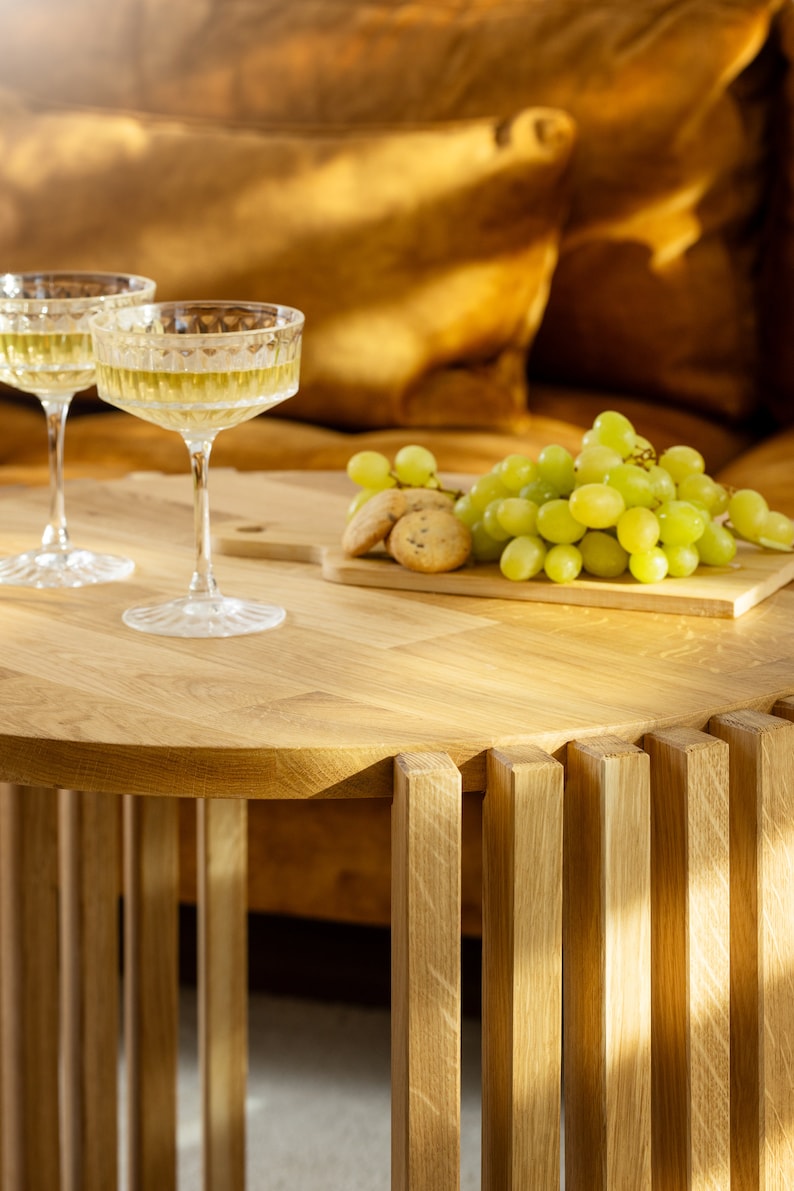 May include: Two glasses of white wine on a wooden table with a bowl of green grapes and cookies. The table has a unique design with vertical wooden slats.