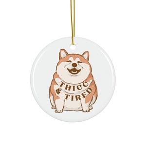 May include: A round, white ceramic ornament featuring a cartoon Shiba Inu dog with brown and white fur. The dog has a smiling expression and the words "THICC & TIRED" are printed on its chest. A gold string is attached for hanging.