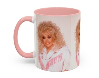 Puede incluir: Una taza de cerámica con asa y borde interior de color rosa claro. El exterior presenta un estampado fotográfico repetido de una persona con voluminoso cabello rubio que viste una camisa blanca con flecos y detalles en rosa.