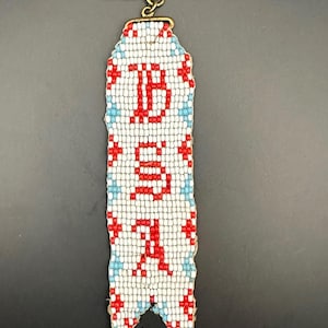May include: A beaded keychain with a blue, white, and red design. The keychain is shaped like a banner and has the word "BSA" spelled out in red beads.