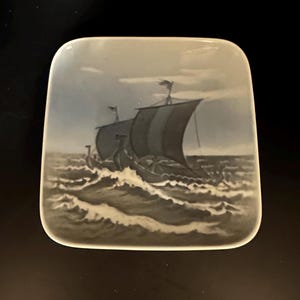 May include: A square ceramic plate with a blue and white image of a Viking ship sailing on a stormy sea.
